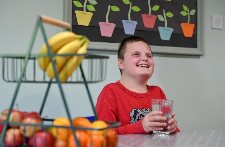 A happy student laughs whilst holding a glass of water sitting next to a fruit bowel - in the background there are paper cut-outs of plants growing stuck to the wall