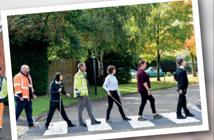 A playful recreation of the iconic Abbey Road crossing scene, featuring six individuals walking in a line on a zebra crossing. The group includes students and staff from New College Worcester, some using white canes for visual impairment. Two adults in high-visibility jackets walk alongside the students. The background features lush greenery and autumn-colored trees, creating a warm and welcoming outdoor setting.
