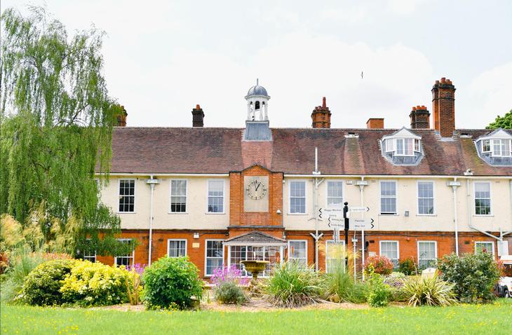 A historic two-story building with a red brick and cream facade, large windows, and a central clock tower, surrounded by a well-maintained garden with colorful flowers and greenery. The scene is bright and summery. 