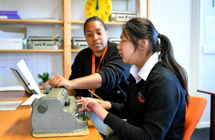 A young woman uses a Braille typewriter while a teacher or support worker sits beside her, guiding her with a hand gesture. They are in a classroom setting with shelves, a large yellow clock, and educational materials in the background.