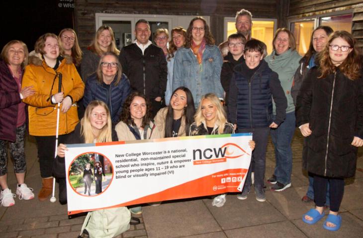 A group photo of smiling people gathered outside a building at night. The group includes young people and adults, some of whom appear to be visually impaired. Four young women are kneeling at the front, holding a banner that reads: 